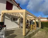 Construction d'une charpente sans ferme en bois, avec des poteaux et des poutres, sur une terrasse. Des échelles sont présentes.