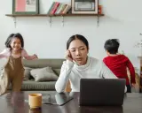 Donna esausta davanti al laptop, mentre i bambini giocano. Un quadro di **parental burnout** in casa.