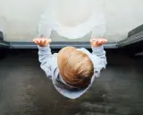 Dziecko w szarej bluzie z kapturem stoi na głowie, patrząc w dół na swoje odbicie.