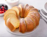 Pokrojona babka piaskowa, oprószona cukrem pudrem, z miseczką owoców leśnych w tle.