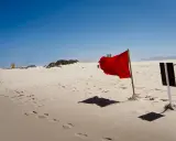 Czerwone flagi u faceta na plaży ostrzegają przed niebezpiecznymi falami. Piasek i błękitne niebo tworzą pustynny krajobraz.