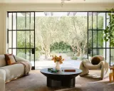 Salones modernos con sofá curvo, sillón acogedor y mesa de centro, conectados a un jardín soleado con olivos.