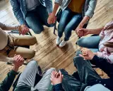 Grupa osób trzyma się za ręce w kręgu, wspierając się nawzajem. Widać tu pracę specjalisty od uzależnień.