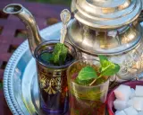 Zwei Gläser mit Minztee, garniert mit frischen Minzblättern, stehen neben einem traditionellen Teekessel und Zuckerwürfeln. Verschiedene Minze Arten verfeinern das Getränk.