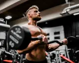 Mężczyzna ćwiczy biceps z ciężarem, wiedząc, że shake proteinowy co daje dla mięśni.