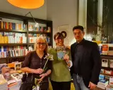 Tre persone sorridono in una libreria, una tiene in mano il libro "Cocco e Magella". Libri in ordine cronologico sugli scaffali.