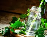 Szklana butelka z wodą i zielono-białą słomką, otoczona gałązkami brzozy z młodymi pąkami. Naturalny syrop z brzozy na kaszel.