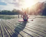 En kvinna med ryggen mot kameran sitter i meditationsställning på en brygga vid en sjö. Solen strålar genom dimman och skapar en magisk stämning. Hon verkar vara helt i nuet.
