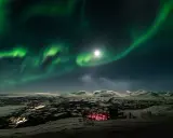 Norrsken över ett snöigt landskap med en upplyst byggnad. Kanske en vy från ett tåg till Abisko.