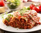 Pyszne cannelloni z mięsem mielonym, polane sosem pomidorowym i posypane parmezanem, z dodatkiem świeżej natki pietruszki.