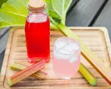 En flaska med röd rabarbersaft och ett glas med is och rosa rabarbersaft står på en träbricka. Rabarberstjälkar ligger bredvid.