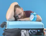 Zmęczony podróżnik śpi na walizce, budzik obok sygnalizuje jetlag.