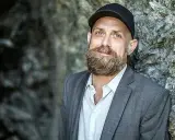 Man med skägg och keps, i grå kavaj, står mot en stenvägg. Kanske en del av en förintelsen serie på SVT.