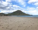 Weite Sandfläche am Playa de los Genoveses mit sanften Wellen und einem grünen Berg im Hintergrund unter einem bewölkten Himmel.