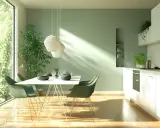 Salle à manger lumineuse avec un mur vert sauge, une grande fenêtre donnant sur la verdure, une table dressée et une cuisine moderne.