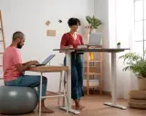 Un homme travaille sur son ordinateur, assis sur un ballon de gym, tandis qu'une femme travaille debout à un bureau réglable. Un espace de travail moderne et dynamique.