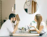 Hombre con personalidad narcisista, frustrado, se cubre la cara con un libro mientras una mujer le habla con exasperación.