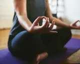 En person sitter i meditationsställning på en lila matta. Händerna vilar i knät med tumme och pekfinger sammanfogade, en vanlig gest för att visa hur man mediterar.
