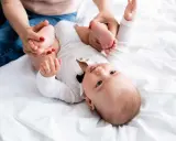 Mama delikatnie ćwiczy nóżki niemowlaka, pomagając mu przy wzmożonym napięciu mięśniowym. Dziecko leży na białym tle.