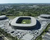 Widok z lotu ptaka na nowoczesny kompleks sportowy, w tym stadion piłkarski i dwie hale widowiskowe. To arena Sosnowiec, otoczona zielenią i parkingami.