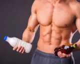 Muskulöser Mann hält Milch und eine Flasche mit bernsteinfarbener Flüssigkeit. Die Frage: Alkohol und Muskelaufbau – eine gute Kombination?