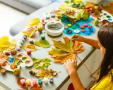 Dziecko tworzy jesienne dekoracje diy, malując liście farbami. Na stole leżą kolorowe farbki, pędzle, liście i kasztany.
