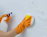 Manos con guantes naranjas limpian paredes blancas con spray y esponja. Guía práctica: como limpiar paredes blancas.