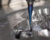 Fresa blu lavora un pezzo metallico, creando dettagli precisi. La cam meccanica è in azione, con liquido refrigerante che schizza.