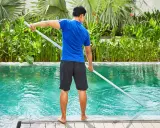 Hombre con camiseta azul y pantalones cortos negros limpia piscina de sal con un limpiafondos.