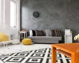Nowoczesny salon z szarą ścianą, na której widać ciekawe efekty. Kanapa, biały fotel i geometryczny dywan tworzą przytulną przestrzeń.