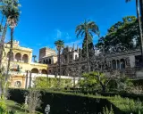 Sonniger Tag im Alcázar von Sevilla. Palmen ragen über kunstvoll verzierte Gebäude und gepflegte Gärten.