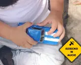 Dziecko bawi się niebieskim autkiem, a obok widnieje żółty znak "Dziecko w aucie", symbolizujący troskę o najbezpieczniejsze foteliki samochodowe.