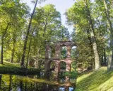 Kamienne ruiny mostu w Arkadii Nieborów, otoczone bujną zielenią i odbijające się w spokojnej wodzie.