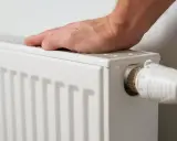 Mano tocando un radiador blanco, listo para cuando encender la calefacción. Termostato visible.