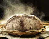 Un pain au seigle rustique, saupoudré de farine, repose dans un panier en osier. La poussière de farine danse dans la lumière, créant une atmosphère magique.