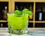 Orzeźwiający drink z likierem miętowym, ozdobiony listkami bazylii, w szklance o geometrycznym kształcie.