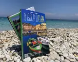 Guida turistica "Isola d'Elba" su una spiaggia di ciottoli, con il mare azzurro sullo sfondo.