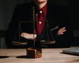 Bilancia della giustizia su un tavolo di legno, con una persona in giacca e camicia rossa sullo sfondo. Le ultime notizie sulla legge Pinto sono in arrivo.
