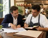 Un asesor de restaurantes revisa gráficos con un chef. Ambos están sentados en una mesa de madera, con el chef tomando notas en un cuaderno.