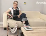 Mujer con overol sentada en sofá beige, sosteniendo una herramienta. A su lado, un revistero diy hecho con cobre y tela.