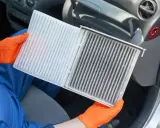 Ręce w pomarańczowych rękawiczkach pokazują brudny filtr kabinowy Toyoty Yaris, gotowy do wymiany.