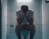 Mężczyzna siedzi na toalecie, ukrywając twarz w dłoniach. W łazience widać rolkę papieru toaletowego. Obraz sugeruje problemy z nietrzymaniem stolca i poszukiwanie domowych sposobów na ulgę.