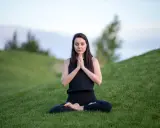 Kvinna i meditationsposition på en grön kulle, med slutna ögon och händerna i bön.