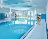 Menschen in Badekleidung am Beckenrand eines Hallenbades mit türkisfarbenem Wasser.