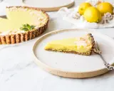 Ein Stück zitronentarte vegan mit Mandelsplittern auf einem Teller. Daneben eine ganze Tarte und frische Zitronen.