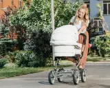 Uśmiechnięta mama spaceruje z białym wózkiem, który jest wyposażony w praktyczne akcesoria.