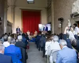 Un evento per il meeting per l'amicizia fra i popoli. Un relatore parla a un pubblico seduto, mentre un altro viene trasmesso su uno schermo.