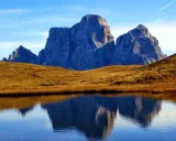 Riflessi maestosi delle Dolomiti nel lago delle Baste, con un prato dorato che separa le acque dalle vette imponenti.