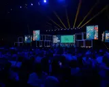 Un grande pubblico partecipa a un meeting per l'amicizia fra i popoli, illuminato da fasci di luce gialla su un palco con schermi.