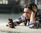 Zmartwiona dziewczyna leży na dywanie, próbując włączyć telefon, który nie chce się włączyć.